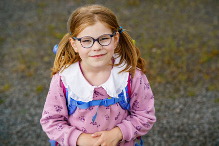 Happy little girl with glasses, wearing a pink sweater and a blue backpack walking to elementary...