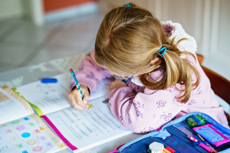 Little girl with glasses, wearing a pink sweater, sits at a table doing homework. Child looks up with a smile, holding a pencil, with an open book in front of her. Indoors, studying, and engaged.の写真素材