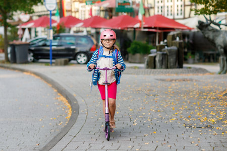 Little happy girl on way to school by pushing scooter. Elementary school child riding in the city, with big satchel. Kid with helmet. Safe route to school and movement for children concept.の写真素材