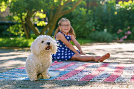 Little girl dressed in a stars and stripes outfit and cute Maltese dog, sits on a paved surface...