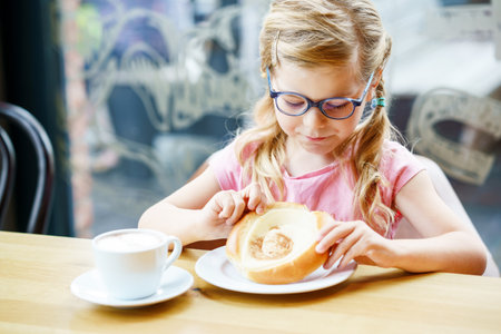 little smiling girl with glasses have a breakfast in a cafe. Preschool child with glasses drinking milk and eating bakery pastry croissant or cake. Happy children, healthy food and meal.の写真素材