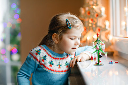 Litte toddler girl sitting by window and decorating small glass Christmas tree with tiny xmas toys. Happy healthy child celebrate family traditional holiday. Adorable baby.の写真素材