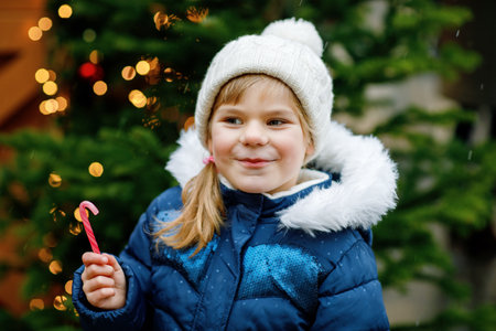 Little cute preschool girl with candy cane from a sweets stand on Christmas market. Happy child on traditional family market in Germany. Preschooler in colorful winter clothes during snowfallの写真素材