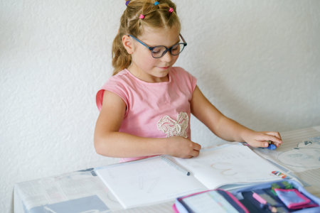 A young girl with glasses, sitting at a table, is focused on writing in her workbook. Child practicing writing numbers with a pencil, engaged in learning. Elementary school, education.の写真素材