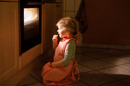 Cute blond girl baking muffins or pie in domestic kitchen. Happy toddler child having fun with helping, sitting near ofen and waiting for cookies. Kid baking for Thanksgiving or Christmas holiday.の写真素材