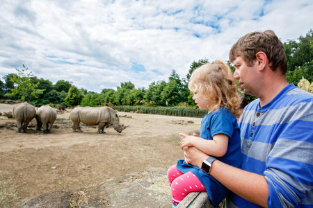 Cute adorable toddler girl and father watching wild rhinos in zoo. Happy baby child, daughter and dad, family having fun together with animals safari park on warm summer day. Irelandの写真素材