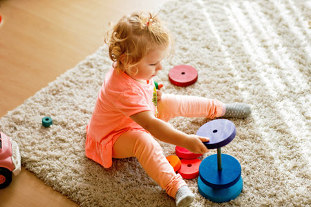 Happy toddler girl sits on a carpet, playing with colorful wooden rings and a toy truck, focused on stacking the pieces. Child plays with rainbow colored pyramid at home or nursery.の写真素材