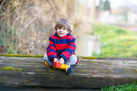 Happy little kid boy in yellow rain boots playing with paper ship boat by huge puddle on spring or autumn dayの写真素材