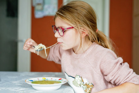 Funny little school girl eat chicken noodles soup with thin pasta. Happy preschool child eating fresh cooked healthy meal with noodles and vegetables at home, indoors.の写真素材