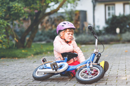 Cute little girl sitting on the ground after falling off her bike. Upset crying preschool child with safe helmet getting hurt while riding a bicycle. Active family leisure with kids.の写真素材