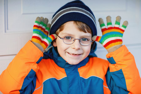 Portrait of a child wearing glasses and colorful winter fashion clothes. Showing a cheerful expression while wearing a blue and orange jacket, striped beanie, and colorful gloves. Winter, spring or autumn fashion.の写真素材