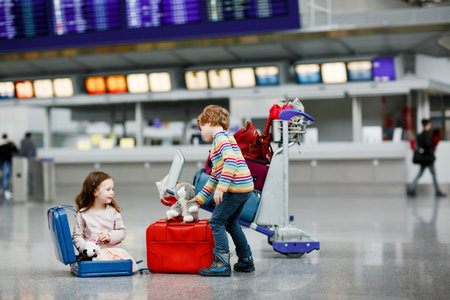 Cute little girl and boy at airport. Tired children, siblings sitting on ground and waiting for delayed flight. Kids playing together by luggage trolley with suitcases at terminal. Travel lifestyleの写真素材