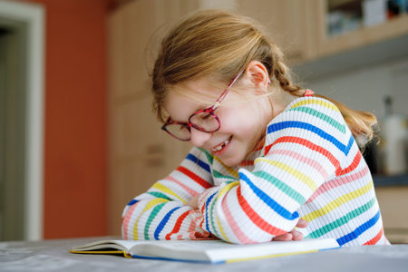 Little happy girl with eyeglasses reading book at home. Elementary school child reads, making...