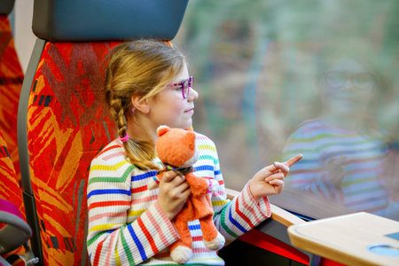 Happy little girl with glasses and a colorful striped sweater sitting by a train window, holding a...