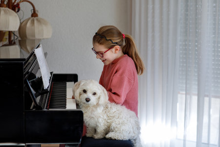 Little happy girl playing piano in living room. Small Maltese puppy dog sitting nearby. Cute preschool child with eye glasses having fun with learning to play music instrument.の写真素材