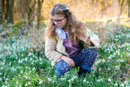 Cute little school girl with eye glasses gathering snowdrop flowers on field in park or forest on a...