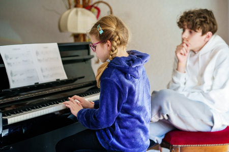 Young little school girl practicing piano while a teenage boy listens attentively. Musical learning, childhood creativity, and concentration. Brother teaching sister playing pianoの写真素材