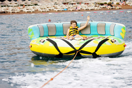 Young boy exudes joy while riding an inflatable tube towed by a boat in the ocean. Happy school child having fun in adventure water park.の写真素材