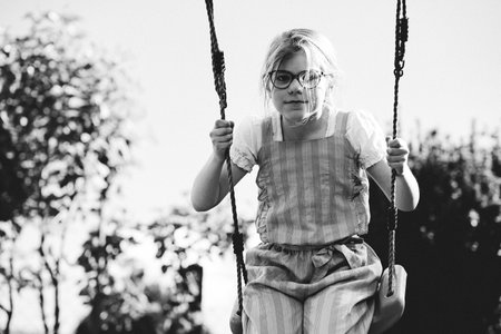 Happy little school girl with eyeglasses having fun on a swing in the garden. Smiling and laughing child enjoying active outdoor play on a sunny day. Childhood joy, motion, and emotions combinedの写真素材