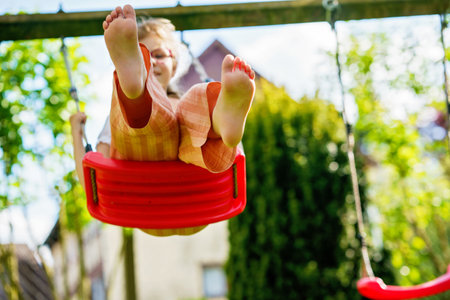 Happy little school girl with eyeglasses having fun on a swing in the garden. Smiling and laughing child enjoying active outdoor play on a sunny day. Childhood joy, motion, and emotions combinedの写真素材