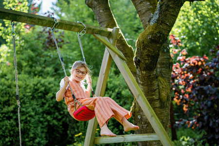 Happy little school girl with eyeglasses having fun on a swing in the garden. Smiling and laughing child enjoying active outdoor play on a sunny day. Childhood joy, motion, and emotions combinedの写真素材