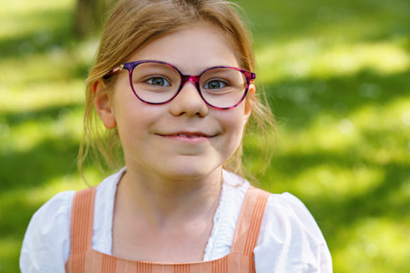Portrait of a happy smiling school girl with eyeglasses in a sunny park. Cute and cheerful child enjoying summer day outdoors. Natural light, childhood joy, and positive emotions.の写真素材