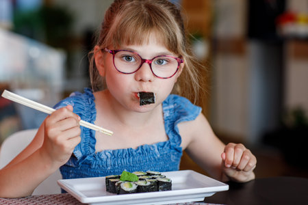 Little school girl with eyeglasses eating sushi with cucumber and avocado in a restaurant. Child using chopsticks. Real life, real people moment. Healthy food and kids nutrition concept.の写真素材