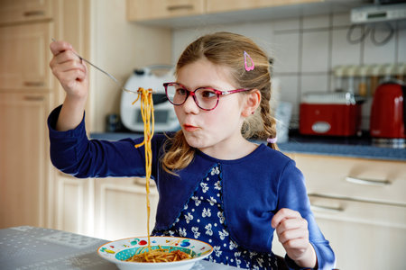 Little school girl eat pasta spaghetti with tomato bolognese with minced meat. Happy preschool child eating fresh cooked healthy meal with noodles and vegetables at home, indoors.の写真素材