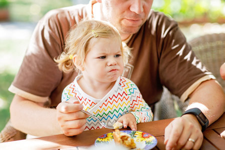 Young middle-aged father feeding cute little toddler girl in restaurant. Kid boy eating on background. Happy healthy family in an outdoor cafe in summer time, eating cakeの写真素材