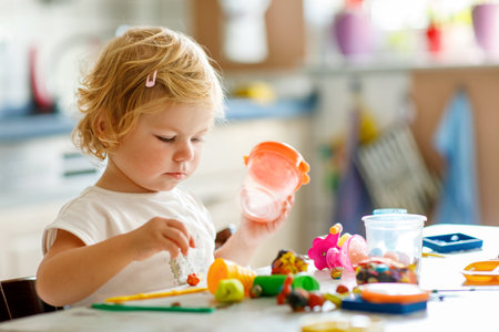 Adorable cute little toddler girl with colorful clay at home. Healthy baby playing and creating toys from play dough at nursery or playschool. Small kid molding modeling clay, creative activityの写真素材