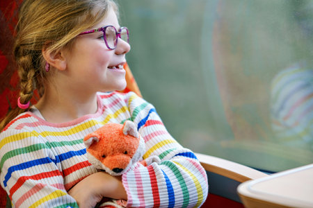 Happy little girl with glasses and a colorful striped sweater sitting by a train window, holding a plush fox toy and looking outside with curiosity. Traveling childの写真素材