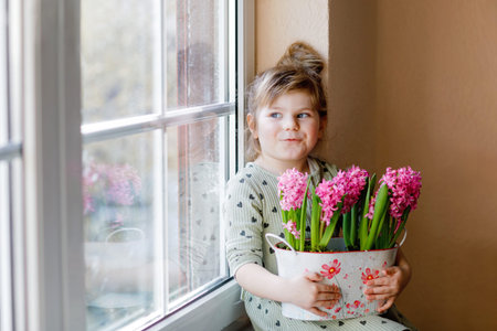Little toddler girl sitting by window with blossoming pink hyacinth flowers. Happy child, indoors. Mothers day, valentines day or birthday and spring concept.の写真素材