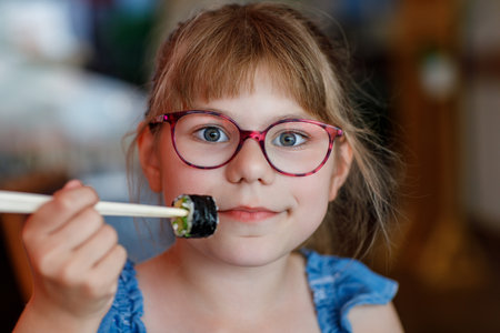 Little school girl with eyeglasses eating sushi with cucumber and avocado in a restaurant. Child using chopsticks. Real life, real people moment. Healthy food and kids nutrition concept.の写真素材