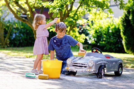 Two happy children washing big old toy car in summer garden, outdoors. Brother boy and little sister toddler girl cleaning car with soap and water, having fun with splashing and playing with sponge.の写真素材