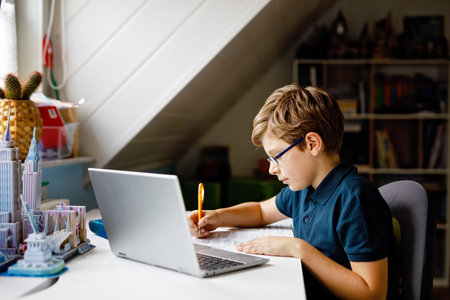 Kid boy with glasses learning at home on laptop for school. Adorable child making homework and using notebook and modern gadgets. Home schooling concept.の写真素材
