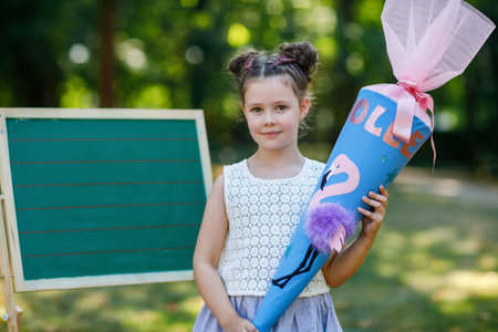 Happy little kid girl by desk with backpack or satchel and big school bag or cone traditional in Germany for the first day of school called Schultuete. Child outdoors. Copyspace on desk.の写真素材