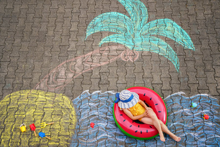Happy little preschool girl in swimsuit on inflatable ring with sea, sand, palm painted with colorful chalks on asphalt. Cute child with having fun with chalk picture. Summer, vacations, summertimeの写真素材