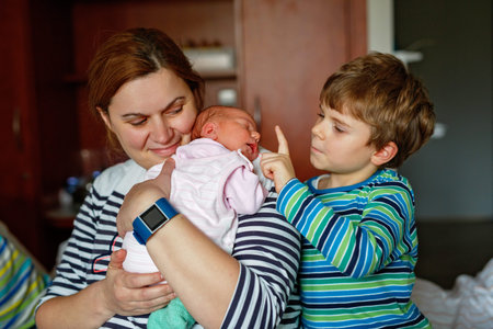 Happy little boy kissing his sleeping newborn sister in hospital bed. Mother holding baby girl on...