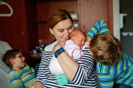 Mother holding newborn baby girl in hospital bed, with two sons sitting beside her. Real life, real people moment of family love, siblings bonding, and joy of welcoming a new child.の写真素材