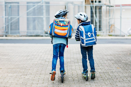Two school kid boys in safety helmet riding with scooter in the city with backpack on sunny day....
