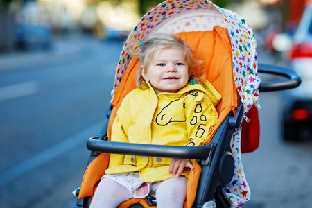 Portrait of little cute toddler girl sitting in stroller or pram and going for a walk. Happy cute...