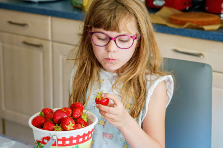 Little school girl with eyeglasses eating fresh sweet strawberries in home kitchen. Healthy food for children. Real life, real people, natural nutrition and fruit snack indoors.の写真素材