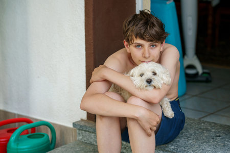 Teenage boy hugging his young Maltese family dog. Real life, real people moment of friendship, love, and bonding between pet and owner. Warm, emotional connection at home.の写真素材