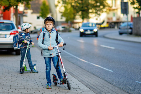 Two school kid boys in safety helmet riding with scooter in the city with backpack on sunny day....
