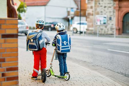 Two school kid boys in safety helmet riding with scooter in the city with backpack on sunny day. Happy children in colorful clothes biking on way to school.の写真素材