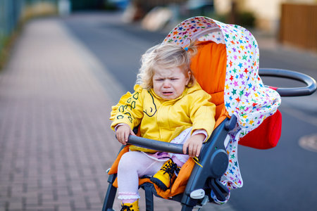 Portrait of little sad toddler girl sitting in stroller and going for a walk. Crying baby child does not want sitting in pram. Healthy daughter. Hysteric crisis of two years phaseの写真素材