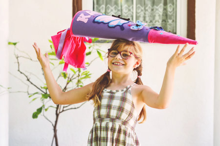 Little girl with eyeglasses and backpack holding a colorful school cone. Happy child smiling, ready...