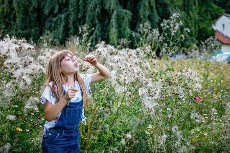 Portrait of happy elementary school girl with glasses playing outdoors in summer meadow, blowing fluffy seeds in nature. Real life authentic childhood moment.の写真素材