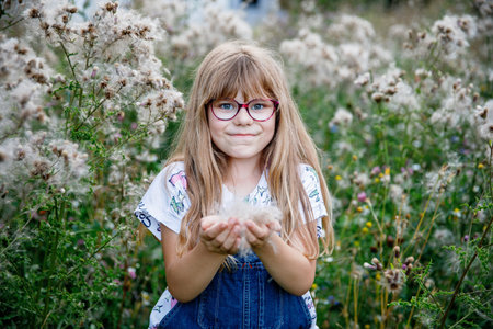 Portrait of happy elementary school girl with glasses playing outdoors in summer meadow, blowing...
