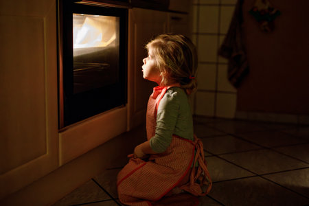 Cute blond girl baking muffins or pie in domestic kitchen. Happy toddler child having fun with helping, sitting near ofen and waiting for cookies. Kid baking for Thanksgiving or Christmas holiday.の写真素材